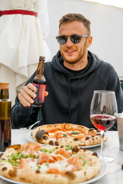 Genç moda eğlence adamı bir bardak şarapla pizza yiyor. Güneş gözlüğü, harika tasasız bir gün, şehir tarzı.