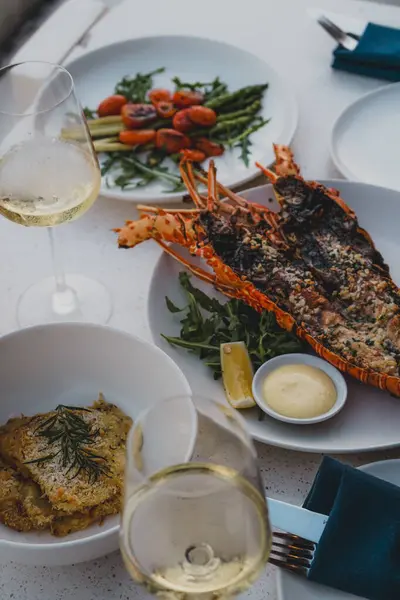 Şık bir sofrada ızgara ıstakoz, kızarmış sebze ve beyaz şarap eşliğinde gurme deniz ürünleri yemeği.