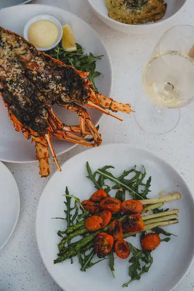 Şık bir sofrada ızgara ıstakoz, kızarmış sebze ve beyaz şarap eşliğinde gurme deniz ürünleri yemeği.