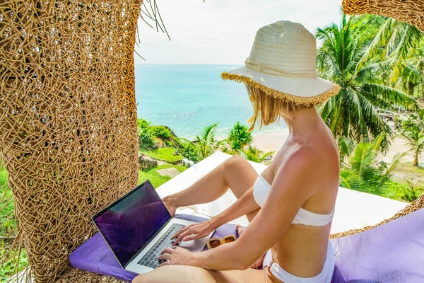 Digital nomad girl working on the tropical beach with laptop computer. Woman in bikini and summer hat using technology for online shopping on vacation. Freelancer.