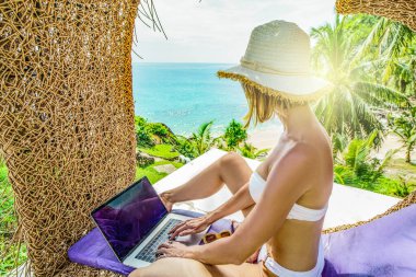 Tropikal plajda bikinili bir kadın bilgisayarla çalışıyor. Dijital göçebe mutlu yaşam tarzı ve teknoloji konsepti. Tayland. 