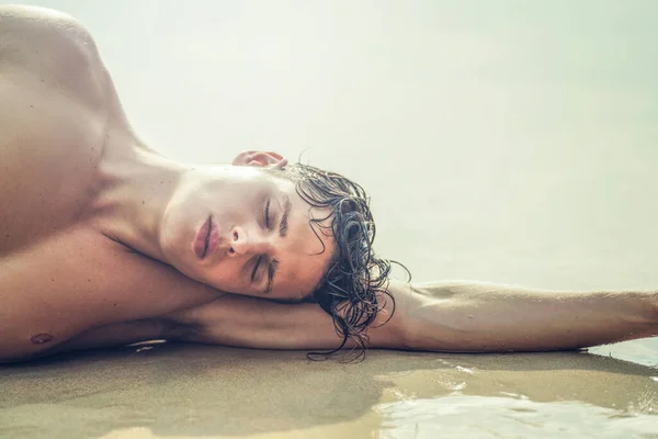 Beauty portrait of handsome young male model relaxing at the beach, lying on the sand. Summer vibes.