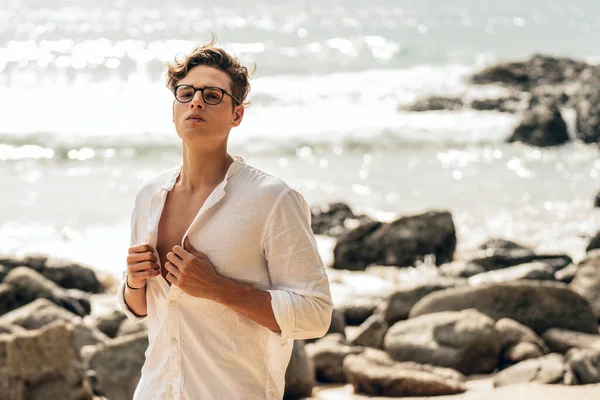 Male model posing in white shirt and eyeglasses on the coast, looking at the camera. Summer vibes. Fashionable young man.