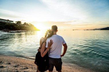 Back view of romantic couple looking at the sunset, relaxing at the beach, enjoying tropical honeymoon. Copy space. Tourism. Thailand. Love
