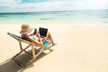 Photo fo a woman in summer hat using mobile phone and laptop computer, working while sitting on a beautiful tropical beach. Digital nomad. Sunny day. 