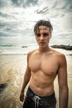 Fit young man posing shirtless on the tropical beach, looking at the camera with smile. Handsome guy relaxing on vacation. Summer time. Traveler. Tropical vibes. Wanderlust.