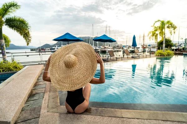Back view of woman wearing big hat and swimsuit relaxing on summer season at resort swimming pool edge. Sunset light. Tourism. Travel.