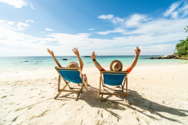 Two happy people having fun on the beach, sitting on blue sunbed with hands raised up, spending leisure time together. Summer holidays concept. Tourism. Travelers