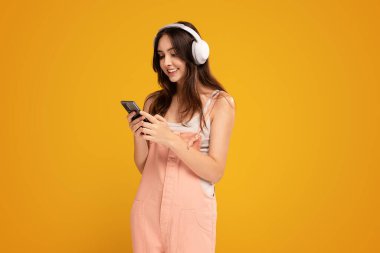Smiling female student listening to music in bluetooth headphones and using smartphone. Studio shot, yellow background, Happy young woman. Online app in mobile phone. Copy space.