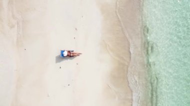 Aerial drone view of one woman in summer hat reading a book on the tropical sunny beach, relaxing. 