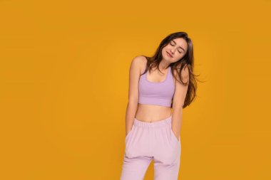 Studio photo of a happy, relaxed, young brunette woman in sporty clothes posing over yellow background with delicate smile and eyes closed. Female student after yoga. Calm, serene girl. A lot of copy space