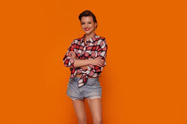 Beautiful young woman with red lipstick looking at the camera, posing in short jeans and fashionable shirt. Orange studio background with copy space.