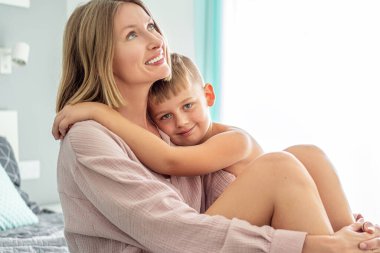 Gülümseyen anne çocuğuyla yatakta oturuyor, sarılıyor, sabah vaktini birlikte geçiriyor. Anne ve oğul arasındaki koşulsuz sevginin şefkatli fotoğrafı.