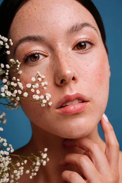Beauty natural woman with freckles, clean and healthy glowing skin. Concept of sunscreen lotions, skincare cosmetics and spa. Girl looking at the camera. Small white flowers on her face. Studio shot