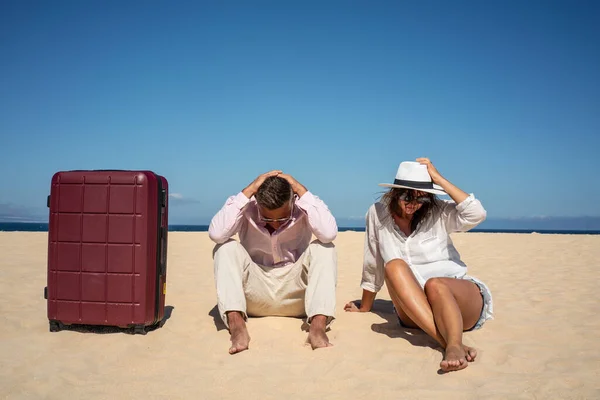 Lost couple of travelers sitting sad on the sandy beach with a big suitcase. Conceptual photo. Copy space. Tourism.