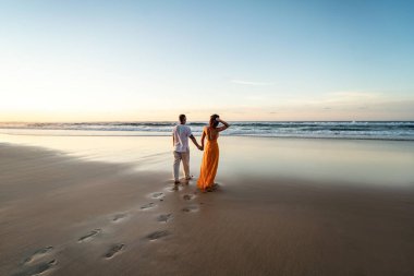 Romantik çift gün batımında sahilde yürüyor, akşam güneşinin tadını çıkarıyor, tropik yaz tatilinde dinleniyor. Balayı. Sevgi. Arkadan bak. Turuncu giysili kadın, keten pantolonlu adam ve beyaz gömlek giyen adam.