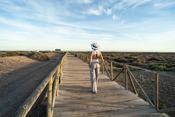 海岸沿いの木製の遊歩道を歩く白い帽子のスタイリッシュな女性は リラックスして晴れた日を楽しんでいます 夏休みだ テキスト用のスペース バックビュー 全長写真 — ストック写真