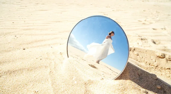 Conceptual photo, reflection in the mirror of attractive blond woman in long white wedding dress posing in desert. Sandy dunes and blue sky.