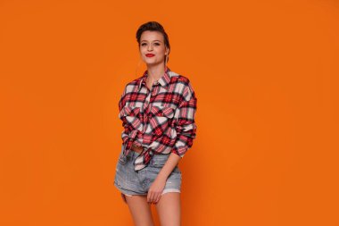Kısa kotlu ve kareli gömlekli mutlu, güzel bir kadının moda fotoğrafı. Turuncu stüdyo arka planında poz veren kız. Bir sürü kopyalama alanı. 