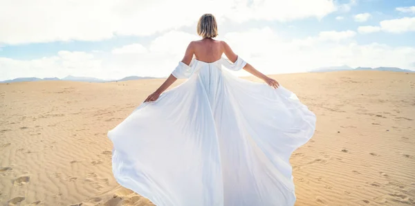 ファッショナブルな マキシ社の白いウェディングドレスは 砂漠の上にポーズをブロンドの女性のバックビューは 日当たりの良い光の中で踊ります 全長写真 スペースのコピー — ストック写真