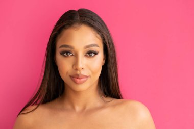 Doğal, güzel, pürüzsüz ciltli ve hassas makyajlı genç bir bayan model kameraya bakıyor, pembe stüdyonun arka planında poz veriyor..