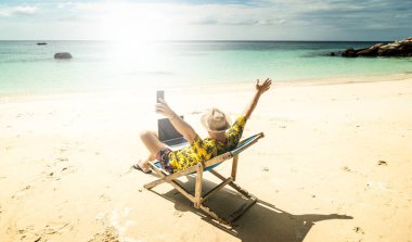 Yazlık şapkalı bir adam, bir iş adamı, dijital göçebe tropik plajda dizüstü bilgisayar ve cep telefonuyla çalışıyor. Serbest çalışan biri. Tropik bölgeler. Yaz titreşimleri