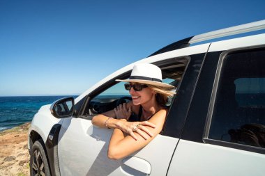 Kiralık arabayla seyahat eden beyaz şapkalı mutlu bir kadın. Macera yaşam tarzı, tatiller. Gülümseyen kadın arabanın camından dışarı bakıyor..