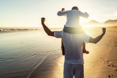 Baba ve oğul, aile tatilinde tropikal plajda boş zamanlarının keyfini çıkarıyor, birlikte oynuyor ve eğleniyorlar. Arkadan bak. Süper kahramanlar. Erkeklerin gücü. Turizm. Gezginler