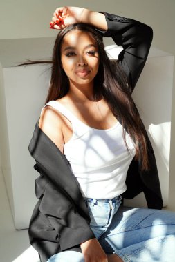 Beyaz atlet tişörtlü, kotlu ve siyah ceketli, stüdyoda poz veren, kameraya bakan neşeli genç bir kadın. Modaya uygun kız.. 