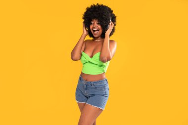 Beyaz kulaklıklı mutlu Afro-Amerikalı kadın yüksek sesle gülüyor, şarkı söylüyor, sarı canlı stüdyo arka planında poz veriyor. Gerçek insanların duyguları. Bir sürü kopyalama alanı.