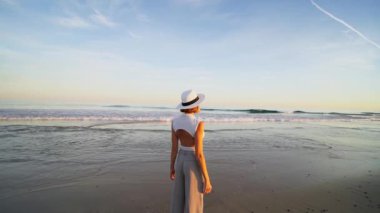 Güzel sahilde duran beyaz şapkalı turist kadının arka manzarası. Gün batımı ışığı. Seyahat konsepti. Hareket eden dalgalar. 