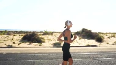 Genç bir kadın sabah antrenmanı sırasında sahil caddesinde koşuyor, koşuyor ve beyaz cep telefonlarıyla müzik dinliyor. Sağlıklı yaşam tarzı kavramı. Sabah rutini.