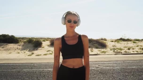 海岸通りに立っている若い女性は 朝のジョギングの後 リラックスして白いハンドホンで音楽を聴いていました 健康的なライフスタイルのコンセプト 朝のルーチン — ストック動画