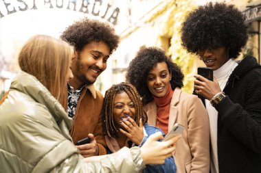 Çok ırklı bir grup genç arkadaş cep telefonundan sosyal medyayı konuşuyor ve izliyorlar. Moda, çok ırklı insanların yaşam tarzı. Açık hava fotoğrafı.