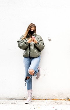 Bir kız cep telefonu kullanıyor, maske takıyor, açık havada mesafeyi koruyor. Kadın cep telefonuna bakıyor ve sosyal medyada yeni içerikler izliyor. 