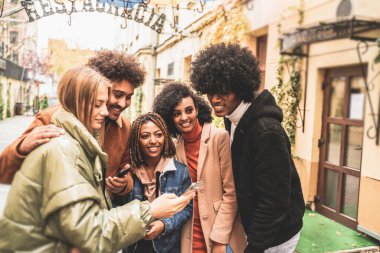 Dışarıda cep telefonuna bakıp gülümseyen bir grup çok ırklı arkadaş. Karışık ırk öğrencileri yeni akıllı telefon teknolojisi kullanarak birlikte eğleniyorlar..
