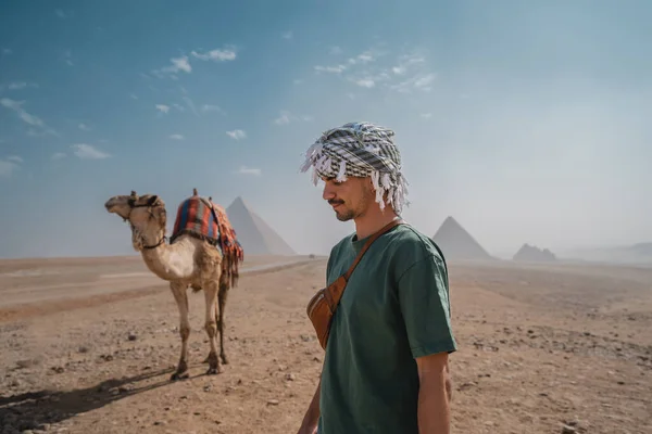 一个戴头巾的年轻人坐在骆驼旁边 后面是金字塔 — 图库照片