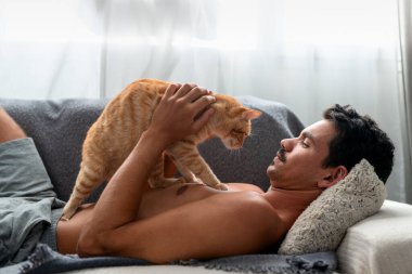 Kanepedeki genç adam kahverengi bir tekir kediyle etkileşime geçiyor.