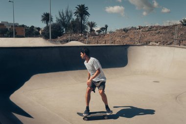 Genç adam güneşli bir günde kaykay parkında paten kayıyor.