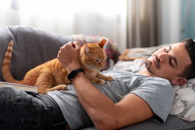  Kanepede uzanmış genç bir adam kahverengi bir evcil kediyle etkileşim halindedir.