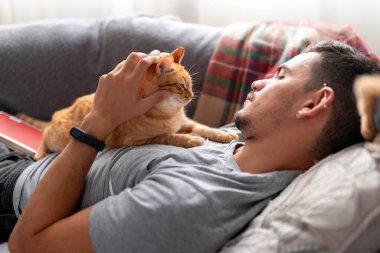  Kanepede uzanmış genç bir adam kahverengi bir evcil kediyle etkileşim halindedir.