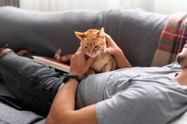  Kanepede uzanmış genç bir adam kahverengi bir evcil kediyle etkileşim halindedir.