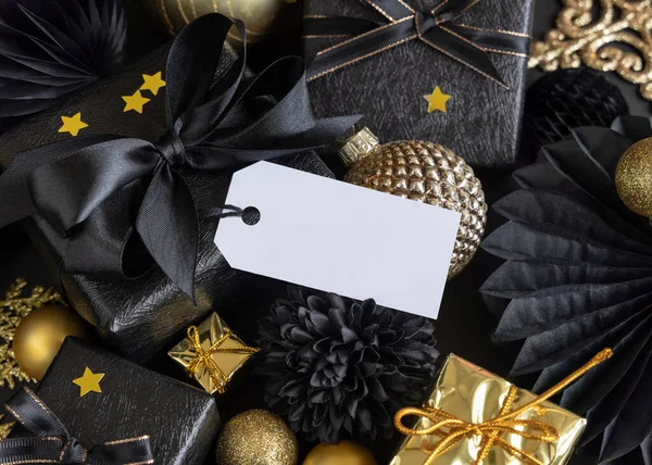 Black and golden Christmas Gift Boxes with a bow and ornaments close up, horizontal paper gift tag mockup, copy space. Dark winter composition with blank label card for Christmas, New Year, Birthday, Anniversary