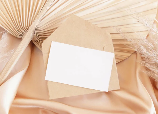 stock image Card and envelope on beige satin fabric and dried palm leaf close up, mockup. Romantic Boho scene with horizontal paper card and envelope. Bohemian or close to nature greeting or wedding Invitatio