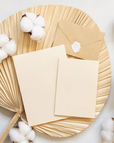 Blank cards and envelope on dried palm leaf with cotton flowers top view, mockup. Romantic scene with vertical paper cards. Bohemian pastel greeting or wedding Invitation