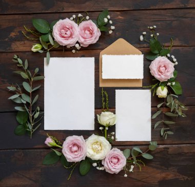 Kahverengi ahşap manzaralı pembe ve krem güller arasında kartlar ve zarf, düğün modeli. Kartlar, zarf ve pastel çiçeklerin olduğu romantik bir sahne. Sevgililer Günü, Bahar veya Anneler Günü konsepti