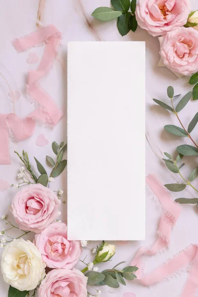 Paper card between light pink roses and silk ribbons on marble top view,  wedding mockup. Romantic scene with vertical blank card and pastel flowers flat lay. Valentines, Spring or Mothers day concept