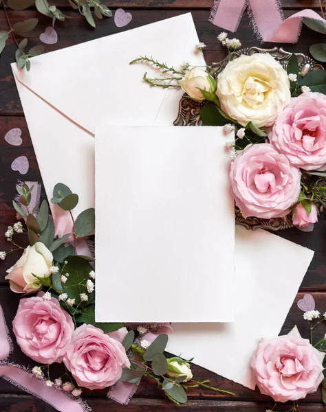 Cards and envelope between pink and cream roses on brown wood top view,  wedding mockup. Romantic scene with cards, envelope and pastel flowers flat lay. Valentines, Spring or Mothers day concept