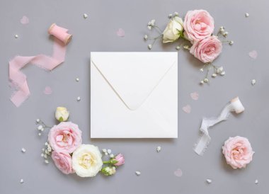 Square envelope between light pink roses and silk ribbons on grey top view,  wedding mockup. Romantic scene with blank envelope and pastel flowers flat lay. Valentines, Spring or Mothers day concept