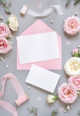 Paper cards between light pink roses and silk ribbons on grey top view,  wedding mockup. Romantic scene with blank vertical card and pastel flowers flat lay.. Valentines, Spring or Mothers day concept